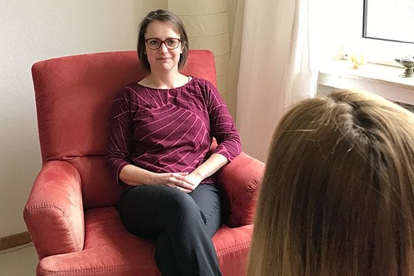 Hier ist ein Foto. Darauf sieht man eine Beratungs-Situation im Frauennotruf Bielefeld e.V. Eine Frau (Beraterin) sitzt in einem roten Sessel und hört freundlich lächelnd zu. Unten rechts im Bild sieht man ein Stück vom Hinterkopf einer zweiten Frau, die beraten wird.